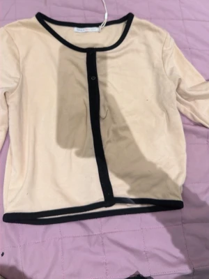 Beige cardigan med svarta kanter - Trendig beige cardigan med svarta kontrastkanter och rund halsringning. Långärmad modell med knappstängning framtill. Perfekt att styla över ett linne eller t-shirt för en clean look.