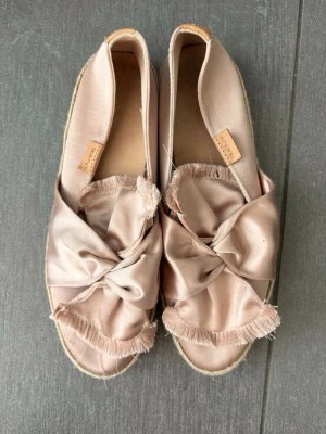 Rosa ballerinaskor i satin från GANT - Säljer ett par ljusrosa ballerinaskor från GANT i glansigt satinmaterial. Skorna har en stor knut framtill med fransiga kanter och en flätad sula i espadrillestil. Perfekta för dig som gillar en feminin och trendig look.