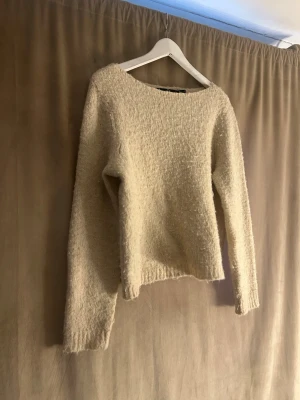 Beige stickad tröja från Vero Moda - Mysig beige stickad tröja från Vero Moda med rund halsringning och lång ärm. Tröjan har en lätt fluffig struktur och är perfekt för att hålla värmen under kyliga dagar. Enkel och stilren design som passar till många olika outfits.