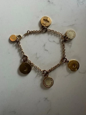 Guldigt charmarmband Marc by Marc Jacobs - Snyggt guldigt armband från Marc by Marc Jacobs med flera runda berlocker. Berlockerna har stjärn- och månformade utskärningar samt ingraverad text. Kedjan är tunn och smidig, perfekt för att ge en cool touch till din outfit.