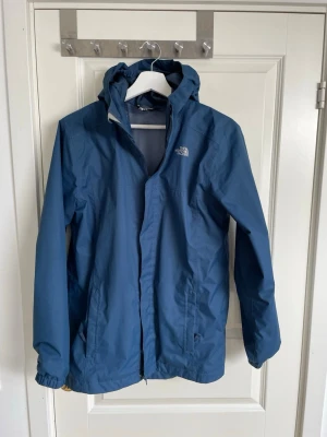 Blå regnjacka från The North Face - Snygg blå XL barn regnjacka från The North Face med huva och dragkedja framtill. Jackan har elastiska muddar vid ärmsluten och två sidofickor. Tillverkad i lätt och vattenavvisande material, perfekt för regniga dagar.