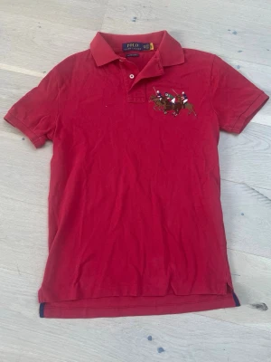 Röd pikétröja från Polo Ralph Lauren - Klassisk röd pikétröja från Polo Ralph Lauren med broderad polospelare på bröstet. Tröjan har korta ärmar, krage och knappar vid halsen. Perfekt till en avslappnad men ändå stilren look.