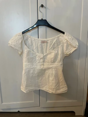 Vit blus med knappar och puffärm - Söt vit blus från Nelly med korta puffärmar och knappar framtill. Blusen har en väldigt liten sminkfläck vid knapparna men man ser den knappast. Annars i bra skick. Pris kan diskuteras. OBS! Köparen står för frakt.