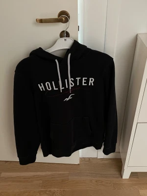 Svart Hollister hoodie med tryck - Svart hoodie från Hollister med vit logga och text på bröstet. Klassisk känguruficka framtill och vita dragsnören i huvan. Mjuk och skön bomullsblandning, perfekt för chill dagar. Oversized känsla och enkel att matcha till jeans eller joggers.