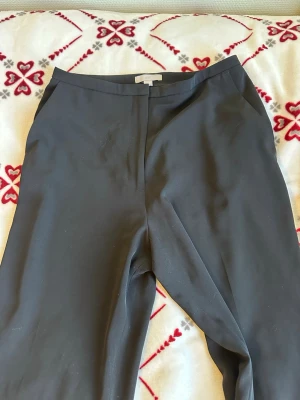 Svarta kostymbyxor med raka ben - Snygga svarta kostymbyxor med raka ben och diskreta sidofickor. Byxorna har en klassisk midja och stängs med dragkedja och hake. Perfekta för en clean och stilren look. Materialet ser ut att vara en mjuk och lätt syntetblandning.