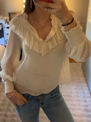 Vit blus från Zara med volang och prickigt mönster  - Söt vit blus med volangdetaljer längs v-ringningen och diskret prickigt mönster. Blusen har transparenta, luftiga långärmar med smock vid ärmslut och axlar.