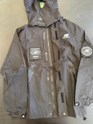 Svart vindjacka från New Balance - Stilren svart vindjacka från New Balance med en sportig design och praktiska detaljer. Perfekt för blåsiga dagar när du vill hålla stilen samtidigt som du är skyddad från vädret.  Detaljer: 	•	Huva för extra skydd 	•	Flera dragkedjeförsedda fickor 	•	Patchdetaljer på bröstet och ärmen 	•	Justerbara muddar 	•	Vita logotyper för en snygg kontrast 	•	Tunt och vindtätt material  Storlek: Passar M–L  Perfekt för både vardag och aktivitet – lätt att matcha med en sportig eller casual outfit.  💬 Hör vi