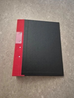 Gaffelpärm - Säljer en klassisk pärm från Jopa med svart omslag och röd rygg. Pärmen har metallmekanism för papper och en etikettficka på ryggen. Perfekt för att organisera dokument eller skolarbeten. Materialet är hård kartong med tygklädd rygg.