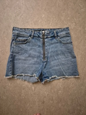 Jeansshorts - Säljer ett par blå denimshorts med hög midja och råa, fransiga benslut. Shortsen har en snygg, synlig dragkedja framtill och klassiska fem fickor. Perfekta för sommaren och ger en avslappnad vibe.