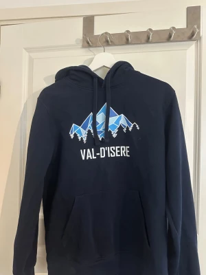 Mörkblå Val d'Isere hoodie - Mörkblå hoodie med tryck av blå berg och texten 'VAL D'ISERE' på bröstet. Klassisk modell med huva och dragsnören samt stor magficka framtill. Tillverkad i mjukt material, perfekt för en avslappnad stil.