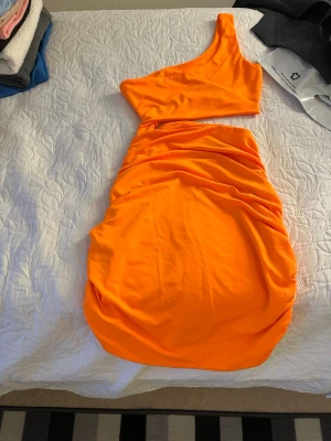 Orange one shoulder klänning - Snygg orange klänning med one shoulder-design och figurnära passform. Klänningen har draperad kjol och är tillverkad i ett stretchigt, mjukt tyg som ger en cool och trendig look. Perfekt för dig som vill sticka ut med färg och form.