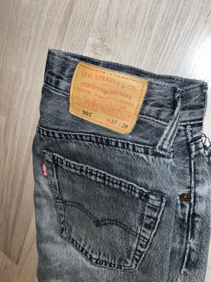 Grå Levi's 501 straight jeans - Säljer ett par grå Levi's 501 straight jeans med klassisk femficksmodell och rå kant vid benslut. Jeansen har en vintage tvätt och en liten slitning på höger ben. Perfekta för en avslappnad och trendig look.