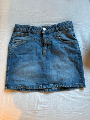 Blå jeanskjol med fickor - Säljer en klassisk blå jeanskjol i rak modell med fem fickor och bälteshällor. Kjolen har knapp- och dragkedjestängning framtill och är gjord i slitstarkt denimtyg. Perfekt att matcha med sneakers eller boots.