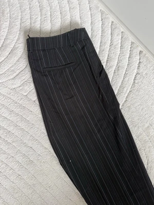 Svarta kostymbyxor  - Snygga svarta kostymbyxor med tunna vita kritstrecksränder. Byxorna har raka ben. Låg midja. Referens: jag är 180 cm (byxorna är precis ovanför marken)