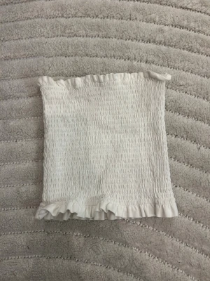 Vit smockad tubtopp med volang - Säljer en vit tubtopp med smockad resår och volangkant upptill och nedtill. Toppen är stretchig och sitter tajt mot kroppen, perfekt för sommaren. Materialet är mjukt och lätt, vilket gör den skön att bära.