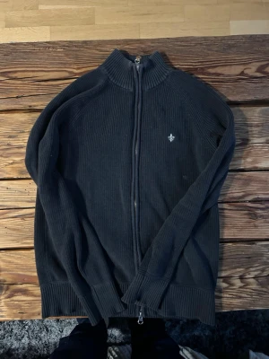 Morris tröja - Säljer en svart ribbstickad ziptröja från Morris med hög krage och dragkedja hela vägen. Tröjan har en liten broderad logga på bröstet och är tillverkad i bomull. Perfekt för dig som gillar stilrena och klassiska plagg. Pris kan diskuteras