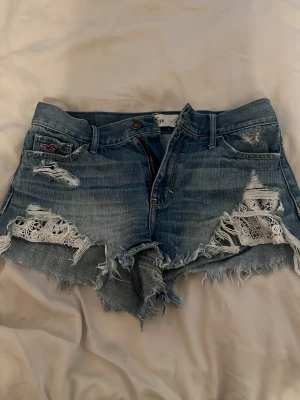 Blå denimshorts från Hollister - Säljer ett par blå denimshorts från Hollister med slitna detaljer och fransig nederkant. Shortsen har spetsinslag i de trasiga partierna framtill och klassiska fickor både fram och bak. Perfekta för sommarens alla äventyr! (Dessa säljs inte längre)