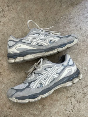 Gråvita Asics sneakers - Snygga sneakers från Asics i grått och vitt med meshpaneler och detaljer i mocka. Skorna har en chunky sula och klassisk Asics-logga på sidan. Perfekta för dig som gillar sportig och trendig stil.