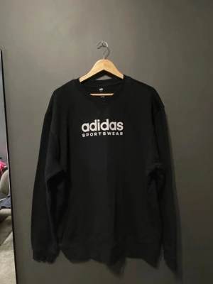 Svart sweatshirt från Adidas - Snygg svart sweatshirt från Adidas med vit logga och texten 'SPORTSWEAR' på bröstet. Tröjan har rund halsringning, ribbade muddar vid ärmslut och nederkant samt en avslappnad passform. Perfekt för en sportig och casual stil.