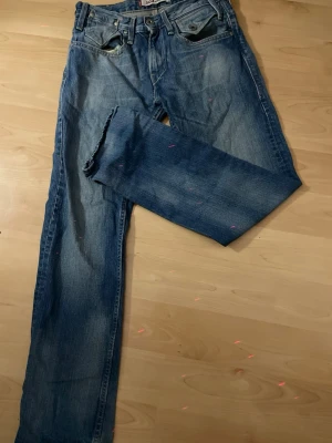 Levi's 504 blå raka jeans - Klassiska blå Levi's 504 jeans med rak passform och fem fickor. Jeansen har slitningar och rå kant vid benslutet för en avslappnad look. Tillverkade i denim med tydliga sömmar och ikoniska bakfickor med V-formad söm.