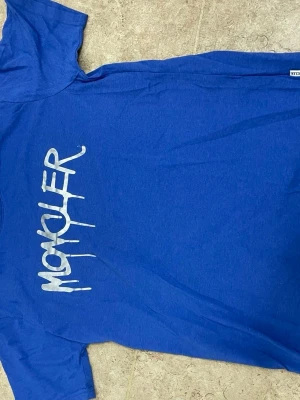 Blå t-shirt med tryck från Moncler - Snygg blå t-shirt från Moncler med vitt grafiskt tryck på bröstet. Klassisk passform och rund halsringning. Perfekt för dig som gillar streetwear och vill ha något enkelt men ändå med en cool detalj.