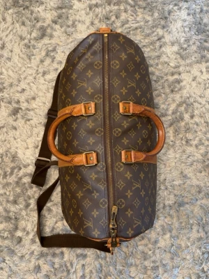 Louis Vuitton KeepallBandoulière 45 - Den ikoniska resväskan för den stilmedvetna resenären. Klassisk Monogram Canvas i tidlöst vintage-utförande med vachettaläder i fin honungsbrun patina. Väskan ar utrustad med axelrem, guldtonade detaljer och "LOUIS VUITTON"-stämplad dragkedja. Tillverkad: Tidigt 1990-tal Skick: 8/10 - fint vintage skick med naturlig patina Nypris: ~25 000 kr Pris: 7290 kr Frakt: Hemlevererat spårbart Äkthet kan verifieras av köparen.