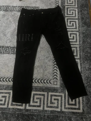Svarta ripped jeans från Amiri - Säljer ett par svarta jeans från Amiri med slitningar och hål på framsidan samt tryckt logga på vänster lår. Jeansen har rak passform och klassisk femficksdesign. Perfekta för dig som gillar streetwear och vill ha en edgy look.