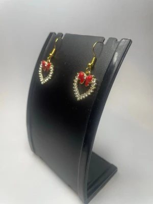 Hjärthängande örhängen med strass - Snygga örhängen med hjärtformad design av små strass och en röd fjäril upptill. Krokarna är guldfärgade och ger en trendig touch. Perfekta för dig som vill sticka ut med söta detaljer och glittrande pärlor.