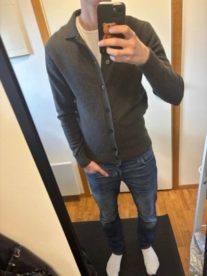  cardigan med knappar - Stilren mörkgrå cardigan med knäppning framtill och klassisk krage. Tröjan har långa ärmar och ribbade muddar vid ärmslut och nederkant. Perfekt att slänga över en t-shirt för en avslappnad men snygg look.