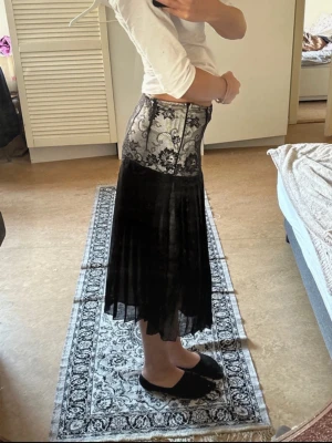 Svart långkjol med spets och plissering - Unik svart långkjol med hög midja och bred spetsdetalj upptill i svart. Kjolen har plisserad nederdel i transparent tyg som ger en cool och elegant look. Perfekt för dig som vill sticka ut med en trendig och feminin stil.