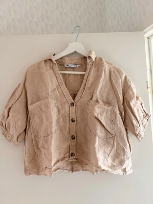 Beige linneskjorta från Zara - Kort beige blus i linne från Zara med avslappnad passform, knappar framtill och två stora bröstfickor. Skjortan har v-ringning och korta ärmar med lätt uppvik.