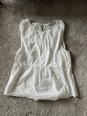 Vit ärmlös blus med broderade detaljer - Söt vit blus i bomull med broderade hål och volang nedtill. Ärmlös modell med rund halsringning och knapp i nacken. Perfekt för sommarens alla tillfällen och ger en fräsch och luftig känsla.