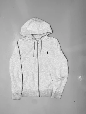 Ljusgrå hoodie från Polo Ralph Lauren - Snygg ljusgrå hoodie med dragkedja från Polo Ralph Lauren. Klassisk design med huva och dragsko, två fickor framtill och den ikoniska lilla loggan broderad på bröstet. Perfekt för en avslappnad och stilren look.