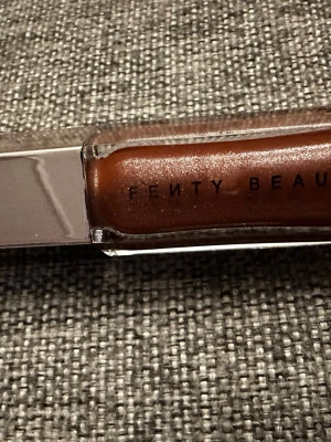 Fenty Beauty brunt läppglans - Snyggt läppglans från Fenty Beauty i en varm brun nyans med skimrande finish. Kommer i en genomskinlig tub med silvrigt lock och tydlig logga. Perfekt för dig som vill ha glansiga och fylliga läppar med en hint av brons.
