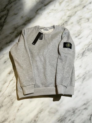 Grå sweatshirt från Stone Island - Grå sweatshirt från Stone Island med rund hals och ribbade muddar. Tröjan har den klassiska svarta patchen med gult och grönt Stone Island-märke på vänster ärm. Tillverkad i mjuk bomullsblandning för en skön känsla och stilren look.