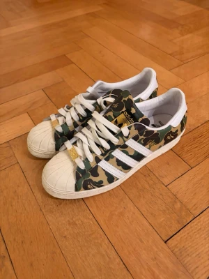 BAPE x Adidas camo sneakers - Unika sneakers från samarbetet mellan BAPE och Adidas med klassiskt grönt camo-mönster, vita ränder och detaljer i guld. Skorna har vit tåhätta i gummi och vita skosnören. Väldigt fint skick, sparsamt använda. Endast lite smuts på framsida tå! Originallådan tillkommer!