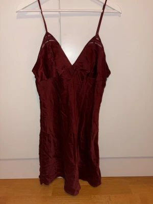 Vinrött nattlinne med spetsdetaljer - Snyggt vinrött nattlinne med tunna axelband och v-ringning. Tillverkat i ett glansigt, mjukt material som känns lyxigt mot huden. Spetsdetaljer vid bysten ger en extra touch. Perfekt för mysiga kvällar hemma. Fint skick. Knappt använd.