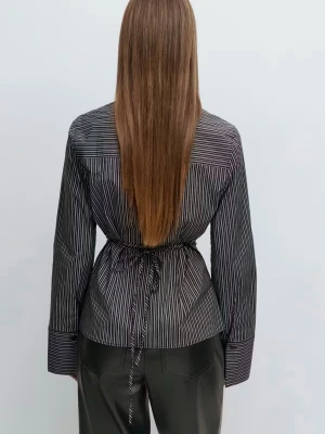 Svart randig skjorta med knytband - Snygg svart skjorta med vita tunna ränder och markerad midja tack vare knytband i ryggen. Skjortan har klassisk krage, långa ärmar med breda manschetter och knäppning framtill. Perfekt för en trendig och stilren look.