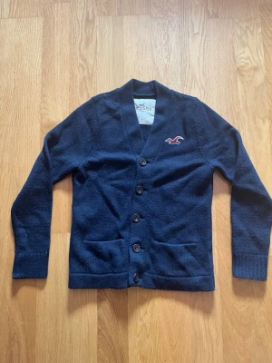 Mörkblå kofta från Hollister - Snygg mörkblå kofta från Hollister med fyra knappar framtill och två fickor. Klassisk logga broderad på bröstet i vitt och rött. Koftan har ribbade muddar och är gjord i mjukt stickat material. Perfekt för lager på lager och chill stil.