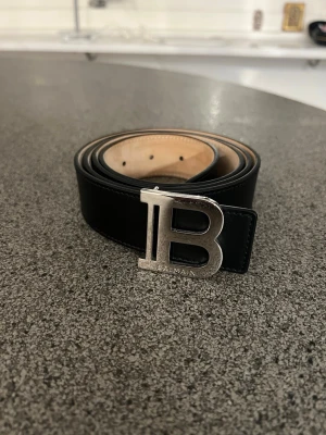 Svart skärp från Balmain med B-spänne Äkta - Säljer ett svart skärp från Balmain med ett stort, silvrigt B-spänne framtill. Skärpet är gjort i slätt skinn och har en stilren look med ljus insida. Perfekt accessoar för att lyfta vilken outfit som helst. Äkta såklart 