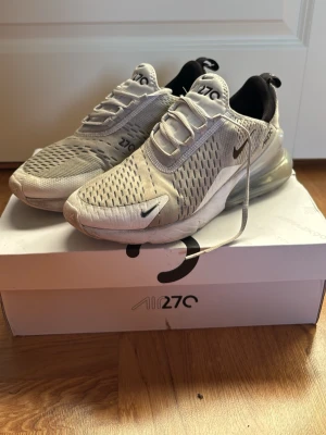 Nike Air Max 270 vita sneakers - Nike Air Max 270 sneakers i vitt med svarta detaljer och stor synlig luftbubbla i hälen. Ovandelen är i mesh för bra andningsförmåga och har klassisk Nike-logga på sidan. Snygg och sportig design som passar till streetwear.