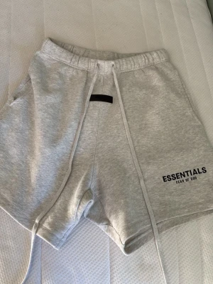 Grå shorts Essentials Fear of God - Snygga ljusgrå shorts från Essentials Fear of God med långa snören i midjan och tryckt logga på ena benet. Mjukt material och elastisk midja gör dem riktigt bekväma. Perfekta för chill dagar eller streetwear-stil.