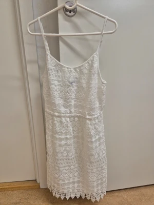 Vit spetsklänning med smala axelband H&M XS  - Söt vit kortklänning med smala axelband och vackert spetsmönster över hela klänningen. Klänningen har en rak passform och spetsdetaljer längst ner vid fållen. Perfekt för sommarens alla tillfällen.