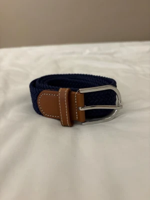 Mörkblått flätat bälte med skinn - Snyggt mörkblått flätat bälte med detaljer i brunt skinn och silverfärgat spänne. Perfekt accessoar för att lyfta din outfit med en klassisk touch. Bältet har vita sömmar på skinnpartierna och är flexibelt tack vare det flätade materialet.