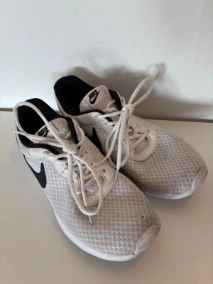 Vita sneakers med svart Nike märke - Säljer ett par vita Nike sneakers med svart nike märke på sidan. Skorna har mesh-överdel för bra ventilation och vita snören. Låg modell med svart innerfoder och klassisk Nike-logga på plösen. Perfekta för en sportig och avslappnad stil. De har använts inomhus och ena skon har ett litet hål vid stortån. Passar snarare storlek 39.