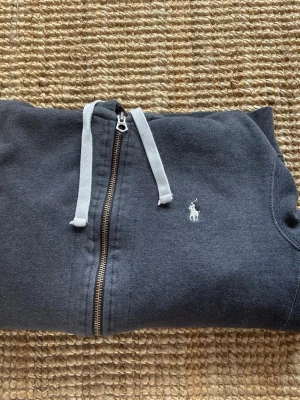 Mörkgrå hoodie från Polo Ralph Lauren - Säljer Dena tvär schyssta  Mörkgrå hoodie ifrån Ralph lauren med dragkedja. Klassisk design med vit broderad logga på bröstet, vita dragsnören och kängurufickor framtill. Tillverkad i mjukt bomullsmaterial och har en skön huva. Ny pris 2295 kr, passar S-M 