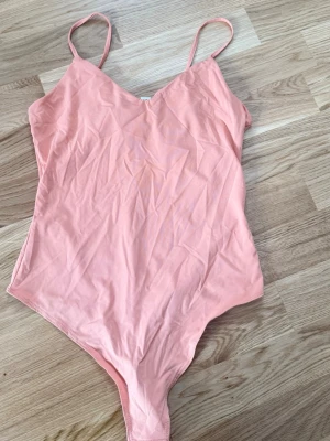 Ljusrosa body med smala axelband - Säljer en enkel och stilren ljusrosa body med smala axelband och v-ringad hals. Bodyn är tillverkad i ett mjukt och stretchigt material, perfekt för att bära under andra plagg. Ingen synlig logga eller märke.