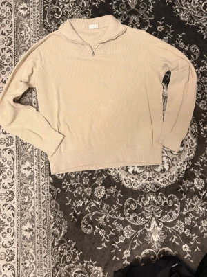  stickad tröja med halvzip  - Snygg beige stickad tröja från Kappahl med halvzip och krage. Tröjan har ribbstickad struktur, långa ärmar och breda muddar vid ärmslut och nederkant. Perfekt för lager-på-lager och enkel att matcha med jeans eller kjol.