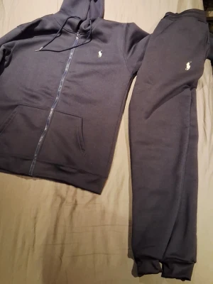 Mörkblått set från Polo Ralph Lauren - Snygg mörkblå hoodie från Polo Ralph Lauren med dragkedja, huva med dragsko och klassisk vit logga på bröstet. Tillverkad i mjukt bomullsmaterial och har en stor magficka framtill. Perfekt för en chill och stilren look.