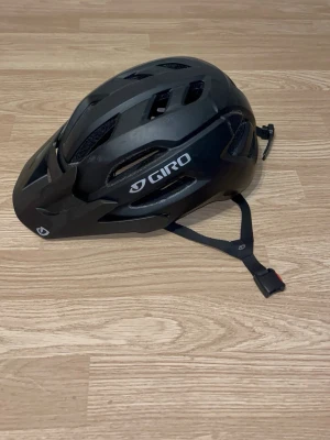 Svart cykelhjälm från Giro - Snygg svart cykelhjälm från Giro med flera ventilationshål och justerbart spänne under hakan. Hjälmen har en sportig design och är tillverkad i lättviktigt material som ger bra komfort och skydd. Perfekt för dig som vill ha stil och säkerhet på cykelturen.